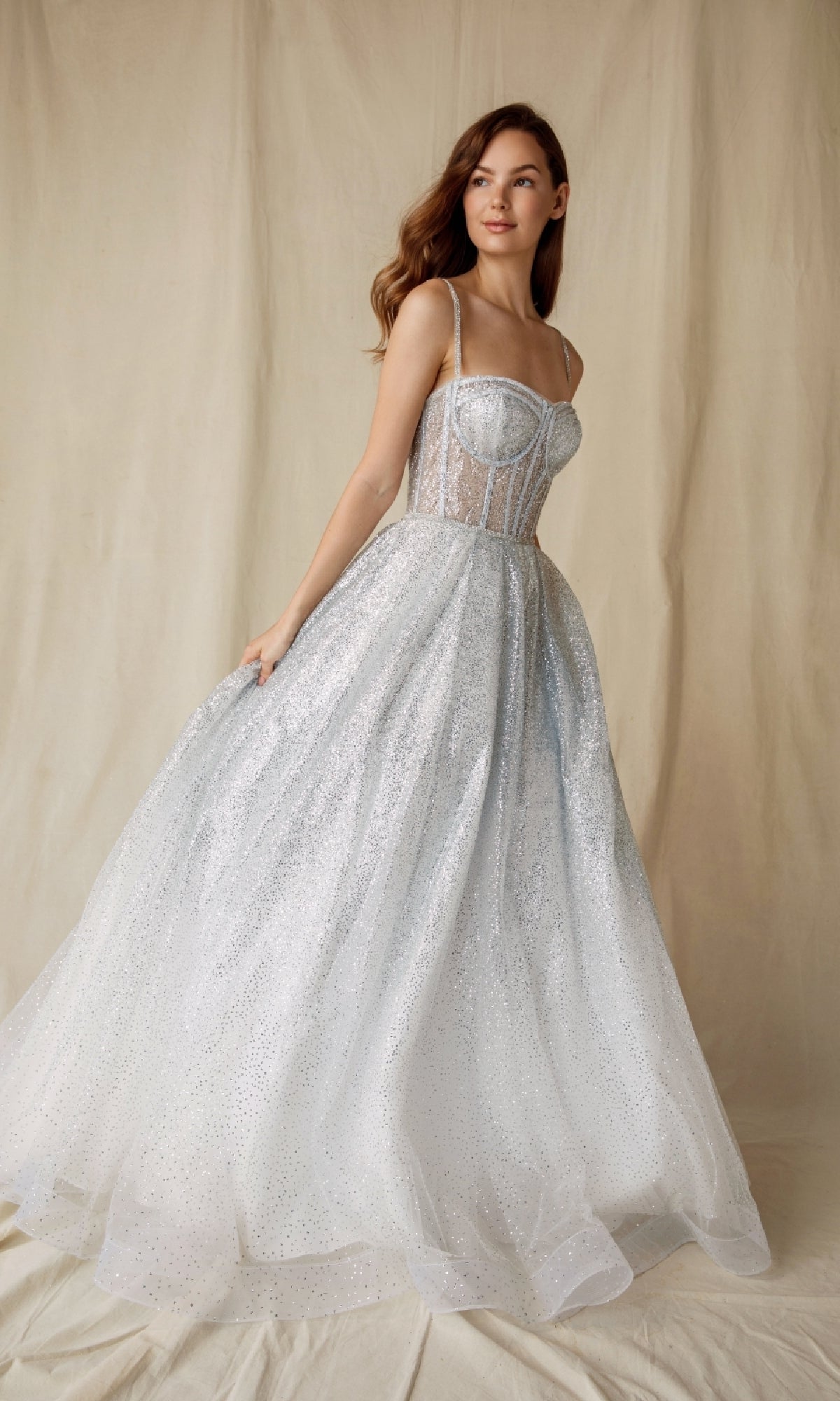 A woman stands before a beige backdrop wearing the Sheer-Corset Glitter Prom Ball Gown: Eureka 9900, a sleeveless white formal gown with a sheer bodice and full, sparkling skirt.